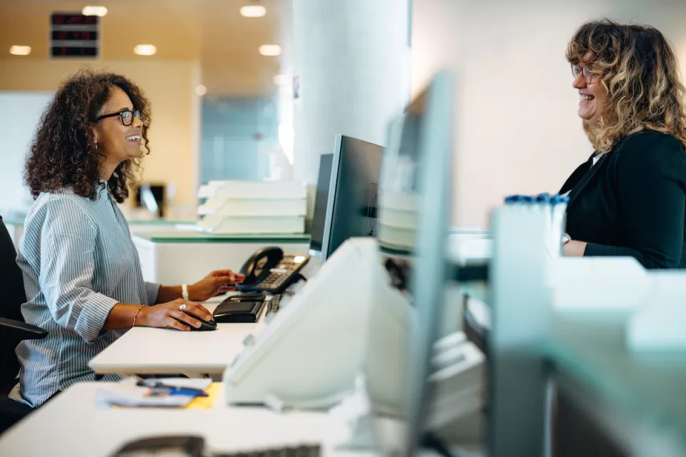 Two women on either side of reception desk talking to each other with row of computers between them.